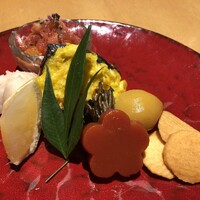 丸新 - [左上から時計廻りで]果物と赤大根のおろし和え,サワラのねぎ焼き,田作り,栗の甘露煮,くわいチップ,人参の羊羹,レモン,百合根の巾着