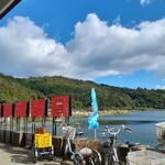 汀の精 - 汀の精さんの前から見た琵琶湖。BIWAKOビエンナーレの作品がある場所に、水色の幟旗があります。赤い看板が並んでいますが、それらが作品。チョークがあり、一枚一枚に質問があり、訪れた人が自由に書きます。