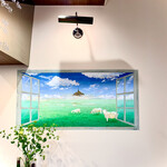 ビストロ ルリール - 窓から眺めるフランスの草原を見事に表現した壁絵。様々な青と緑の色彩が、白くぽっかり浮いた雲を引き立てています。山羊と目が合うのが楽しい