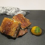 レミニセンス - 鰻：鰻を白焼きにしてから ワインを使用しフレンチテイストに仕上げた三河一色産の鰻は、炭火でパリッと焼きあげられた皮の食感と香ばしさ、ウナギ本来の美味しさが熱々・最高の味わいで堪能できます。鰻の下には、牛のタルタルと 香りづけに棒葉が敷かれ、胡麻・赤ワインのソースが 鰻の美味さを更に引き立てています。 脇には うずら卵の黄身が添えられています。