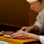 くるますし - ここの大将、炙ったり、湯に透したり、切りつけたりと一人でこなすので、厨房と付け場を行ったり来たりで忙しいのです。 藁の薫香がしてたと思ったら、炙られた大きな切身が登場。 温かいうちに供するため、大将、急いで、静かに切ってます。 僕らは、それをヨダレたらしたワンコにのように待ってます。