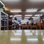 源蔵 - 店内はクリーム色の壁と腰高まで青系灰色の壁紙、赤系統の床シートと淡い色合いのテーブル昔ながらの食堂然とした雰囲気
            お席はテーブル4席×12卓とテーブル2席×5卓の合計58席