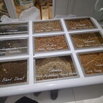 アラビカ東京 麻布台ヒルズB1店 - インドネシア3種、コロンビア5種が生豆で並ぶ。生豆の色も様々。オーダーごとに店頭の焙煎機で好みの焙煎度に仕上げられる。Colombia Quindio Santa Monica は中央奥。