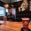 HIROSHIMA KITA BEER THE BASE - ガラス張り壁の向こうには広島駅のバスターミナルと行き交う人々が見え、キッチンエリアの黒塗り壁にはビールを飲む女性のイラスト、12連のビールTapがあり、お洒落なビールスタンドといった趣き、BGMは洋楽