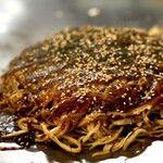 来んさいや - 焼き上がり。 オタフクお好み焼きソースを塗って、ごまと青のりをふりかけます。  まだ鉄板の熱でソースがジュウジュウと焼けてます。 熱いうちに、 口の中を火傷しながらどうぞ！