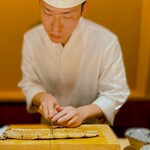 くるますし - 一品目、いきなりきました。 大型の天然うなぎの炙り。 これを8等分していきます。 鰻もデカいけど、大将の顔もデカく撮れちゃった。 ほんとはこんなにデカくないですから（笑）。