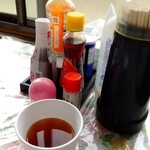 ながおかお惣菜店 - テーブル席上の調味料他達と冷たい麦茶