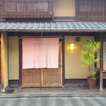 飯田 - 阪急電車烏丸駅から徒歩10分の「飯田」さん
            外観は京町屋が建ち並ぶ風情ある通り、町屋造りの建物、淡い鴬色の土壁、木製引戸扉、桜色の暖簾、趣を醸し出す佇まい