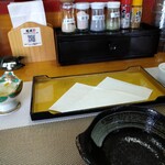 天ぷら定食専門店 起天 - カウンターです。手前の黒い小鉢が天つゆの器。左はお漬物　右は大根おろし。向こうの黒い箱にお箸（割り箸）とおしぼりがありました。その上は定番の各種の調味料。真ん中の金色のお盆に天ぷらが載ります。