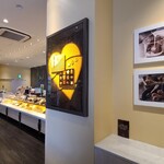 GODIVA - 店内は白い天井、ガラス張り壁&白壁&大きな電灯のブランドマークがあしらわれたカウンター後の濃灰色のタイル貼り壁、濃灰色の木板張り床、高級感のある空間、BGMは洋楽