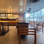 スターバックスコーヒー - 店内は黒塗り天井、ガラス張り壁&壁画のある茶灰色の壁&外壁のガラス張り壁、茶灰色の柱、灰色のタイル貼り床、フードが陳列されたショーケース&雑貨が陳列された木製棚、ゆったりとした空間、BGMは洋楽