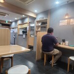 江戸そば 孫吉 - 店内は白い天井、淡いブルーグレーの壁、グレーのタイル貼り床、ナチュラルウッドのカウンター&テーブル、落ち着けて和める空間、BGM代わりにTV音声が流れています
            カウンター席、テーブル席で合計16席
