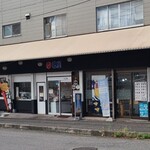 巴屋 - 3階建て建物の1階にあり、クリーム色の防水布の庇、上は黒塗り壁&真ん中は白塗り壁&下は黒塗り壁のストライプ、ガラス窓とガラス張り自動扉、写真入りのイメージパネルが貼られ、親しみが持て入り易い佇まい