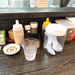 長浜ラーメン三吉 - 卓上も清潔。レンゲを蓋付き容器に入れてるなんて、以前の三吉では考えられない。そーいや、昔はR34はさんで向かいのヨコオ釣具の店の人が調理場に入っていたような記憶が…