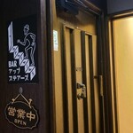 Bar Upstairs - 外観は飲食ビル5階にあり、エレベーターを降りると階段を上がる人物の看板、黒塗りの木製開き扉、重厚さと少しのユーモアを感じる佇まい