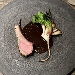 レヴォ - 黒い陶器皿の中央に薪火でローストされた猪肉がしっとりと佇みます。外側は香ばしく焼き色が入り、中心は淡く赤みを残した火入れ。