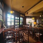 バン - 店内は黄灰色の天井、ガラス張り壁&茶色の柄付き壁、赤茶色の防水シート貼り床、赤紫色な天板テーブル、藤の椅子、茶系の調度品、小さなキッチンでコンパクトな空間、BGMはクラシック系のラジオ音声