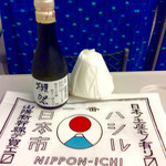 Jeiaru Nishinihon Fudo Sabisu Netto - 「獺祭 大吟醸 精米率35%」(180ml瓶850円)。横の白いのは紙製のぐい呑み(包装状態)。お土産用の袋も貰える。